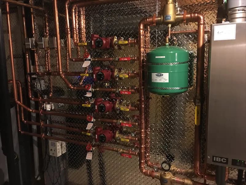 Copper hydronic manifold with four red circulator pumps and a green expansion tank mounted above a condensing boiler